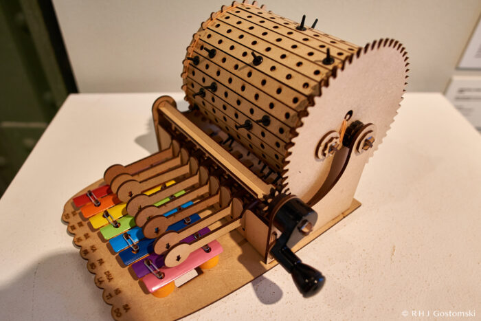 Palacio de Cibeles, Madrid A wooden mechanical music box with a hand crank, colour-coded metal keys, and a rotating drum with pegs that strike the keys to produce musical notes—part of a modern art display at Palacio de Cibeles, Madrid.