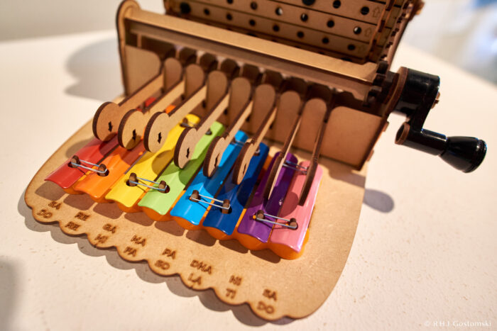 Palacio de Cibeles, Madrid A close-up of a colourful wooden xylophone with rainbow-coloured keys, each labelled with a musical note. A black handle is visible on the side, suggesting it is a mechanical musical instrument.