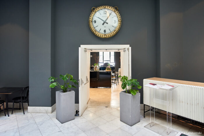 Palacio de Cibeles, Madrid A large clock hangs above open double doors leading to a bright café area. Two potted plants flank the entrance. Black chairs and tables are on the left, and a radiator with a menu stand is on the right.