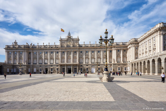 The image shows the grand façade of The Royal Palace in Madrid, with a Spanish flag atop the building, ornate street lamps, and people walking across the spacious stone courtyard under a partly cloudy sky.