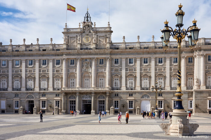The Royal Palace, Madrid The Royal Palace of Madrid with a Spanish flag flying atop, ornate lamp-posts in front, and people walking across the large plaza under a partly cloudy sky.
