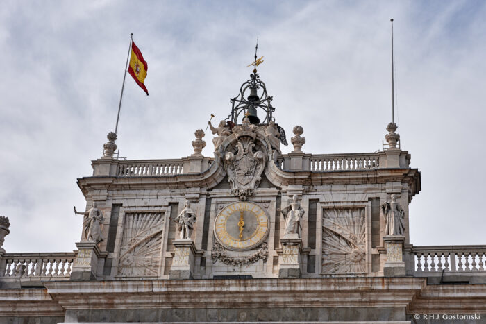 The Royal Palace, Madrid The ornate façade of a historic building with a clock, statues, and Spanish flags on top. The sky is cloudy in the background.