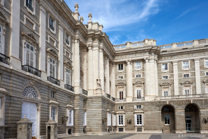 The image shows the exterior of the grand, ornate Royal Palace in Madrid, featuring tall columns, arched windows, and detailed stonework under a bright blue sky.