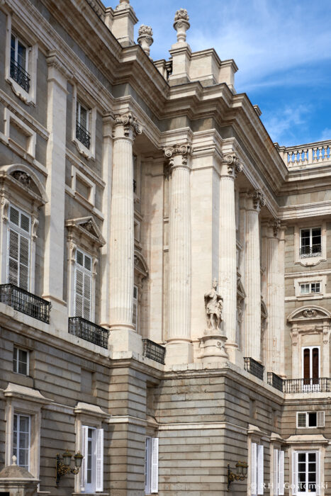 The Royal Palace, Madrid A grand neoclassical building façade with tall Corinthian columns, ornate stone carvings, balconies with black railings, large windows, and a clear blue sky in the background.