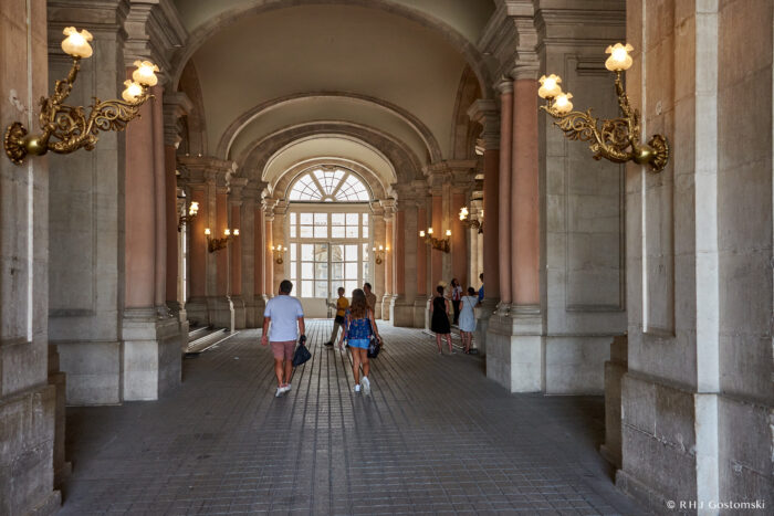 The Royal Palace, Madrid People walk through a grand, arched hallway with tall columns, ornate wall lamps, and large windows letting in natural light. The scene has a historic, elegant architectural style.