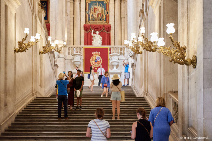 Inside the Royal Palace in Madrid, tourists walk up a grand staircase lined with ornate golden lamps. At the top, a large red and gold royal crest sits beneath a statue, whilst visitors admire the decor and take photos.