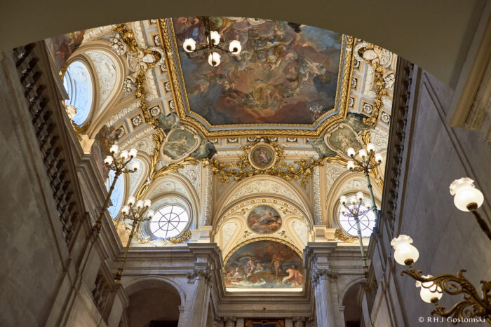Ornate ceiling of a grand hall in the royal palace, Madrid, featuring elaborate gold detailing, round windows, and detailed frescoes with classical scenes—like Giaquinto’s fresco above the staircase—illuminated by elegant chandeliers.