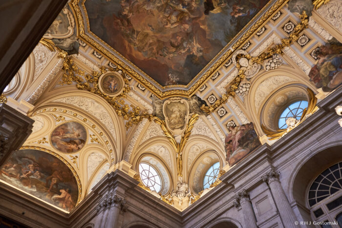 The Royal Palace, Madrid Ornate, baroque-style ceiling with detailed frescoes, gold accents, and arched windows letting in natural light; intricate architectural and artistic elements are visible throughout.