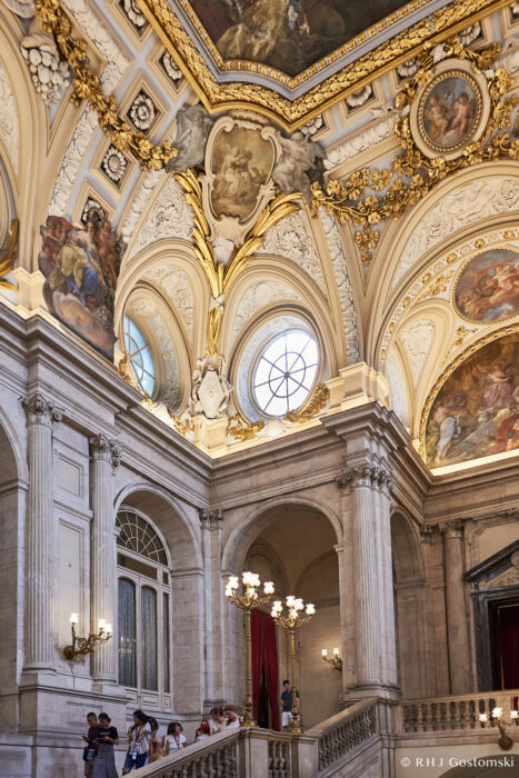 The Royal Palace, Madrid Visitors stand on a grand staircase inside an ornate palace, featuring marble columns, gold-trimmed arches, large round windows, and detailed frescoes on the vaulted ceiling. Lush red curtains hang beside the stairs.