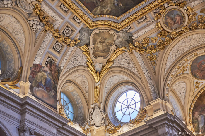 The Royal Palace, Madrid Ornate baroque ceiling with gold accents, elaborate plasterwork, circular windows, and detailed paintings depicting classical scenes and figures. The rich architectural details create a grand, historic, and elegant atmosphere.