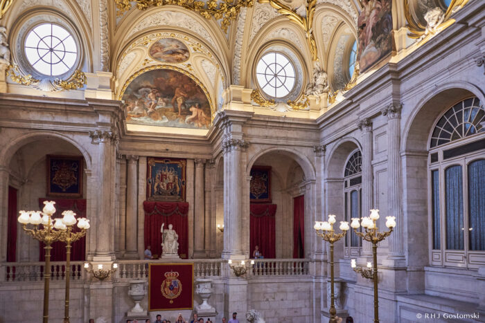 The Royal Palace, Madrid A grand hall with ornate ceilings, large arched windows, red curtains, chandeliers, and classical statues. The walls are adorned with paintings and a royal crest above the central statue. Visitors look on from balconies.