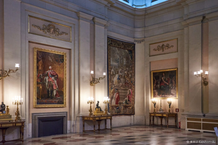 Inside The Royal Palace, Madrid, a grand room boasts ornate wall mouldings, large framed paintings, and a tapestry. Gilded furniture, candelabras, and marble floors complete the elegant, historic atmosphere.