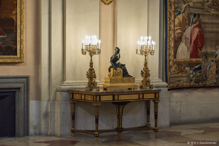 The Royal Palace, Madrid Ornate table with a decorative clock and statue centrepiece, flanked by two gold candelabra with lit candles, set against an elegant wall with tapestries and framed art in a grand interior.