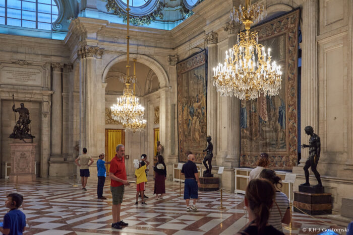 The Royal Palace, Madrid People explore a grand hall with marble floors, chandeliers, statues, and large tapestries on the walls. The room has high ceilings, arched doorways, and is filled with natural light from tall windows.