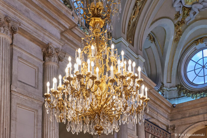 The Royal Palace, Madrid A grand, ornate crystal chandelier with lit bulbs hangs from a high, elaborately decorated ceiling in an elegant room with columns, arches, and a round window.