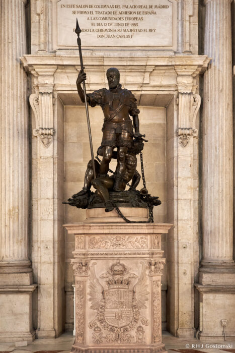 The Royal Palace, Madrid A bronze statue of a knight holding a spear stands atop a decorated plinth in an ornate hall with a stone inscription and columns in the background.