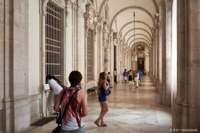 The Royal Palace, Madrid People walk and stand in a long, arched stone corridor with tall columns and barred windows. Some are taking photos or looking around, whilst sunlight illuminates the marble floor.
