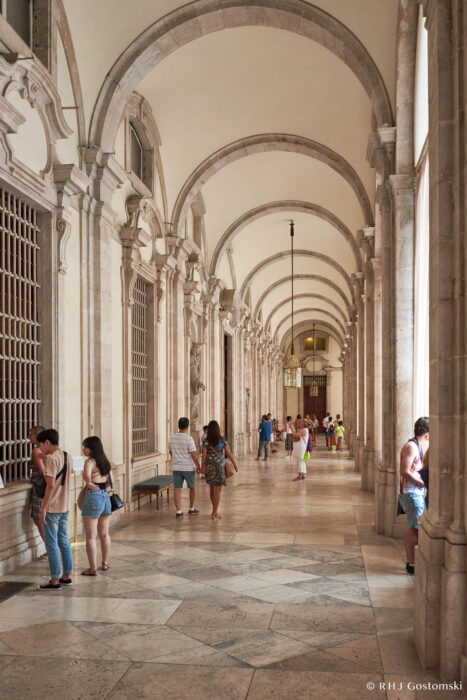The Royal Palace, Madrid People walk and stand inside a grand arched hallway with marble floors and tall columns. The corridor has ornate details, large windows on the left, and warm natural light fills the space.