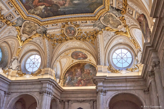 The Royal Palace, Madrid Ornate interior of a grand building with arched windows, intricate gold and white details, and painted ceiling frescoes featuring classical figures and scenes. Marble columns frame the elaborate architectural design.