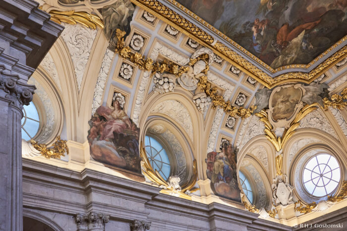 The Royal Palace, Madrid Ornate palace ceiling with golden trim, decorative mouldings, round windows, and detailed frescoes depicting figures in flowing robes beneath painted sky scenes.