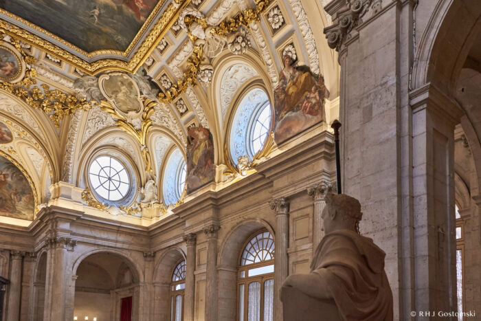 The Royal Palace, Madrid Ornate palace ceiling with golden trim, decorative mouldings, round windows, and detailed frescoes depicting figures in flowing robes beneath painted sky scenes.