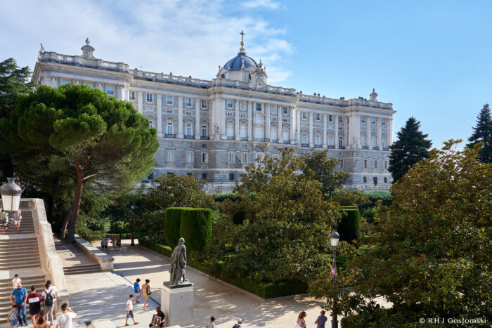 The Royal Palace of Madrid, a grand historic building with ornate architecture, stands behind lush green trees and gardens. Visitors stroll along paths and steps in the foreground, enjoying a sunny day in the heart of Madrid.