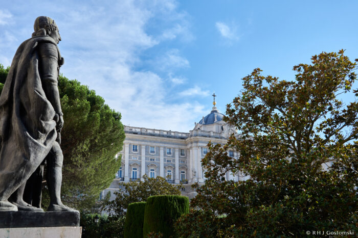 The Royal Palace, Madrid A stone statue of a man stands in a garden with green trees, facing a grand, ornate palace with a blue dome and golden details under a bright blue sky.