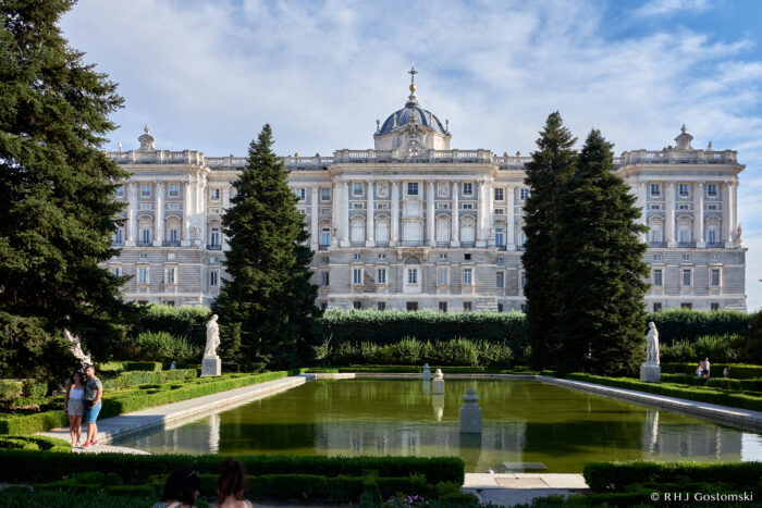 The Royal Palace viewed from the Sabatini Gardens stands behind manicured lawns, a large rectangular pond, tall trees, statues, and a few people enjoying the sunny day.