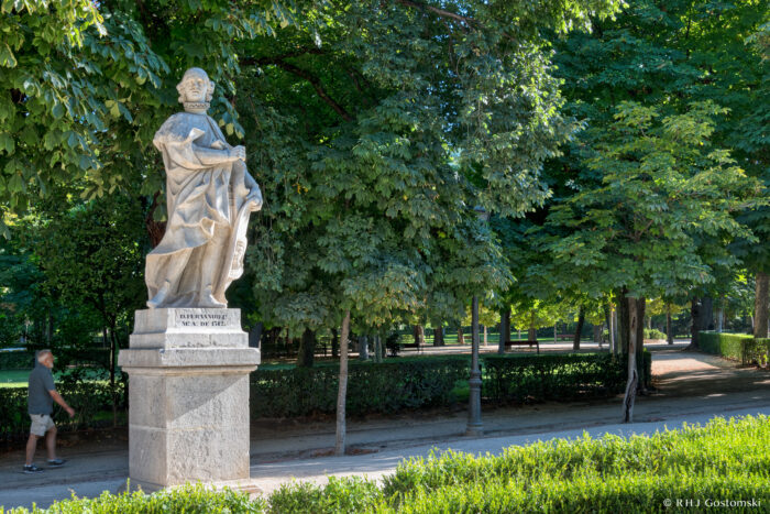 Statue of Fernando IV A stone statue of a robed Fernando IV stands on a plinth in a park surrounded by green trees, with a person walking on a path nearby. Sunlight filters through the foliage.