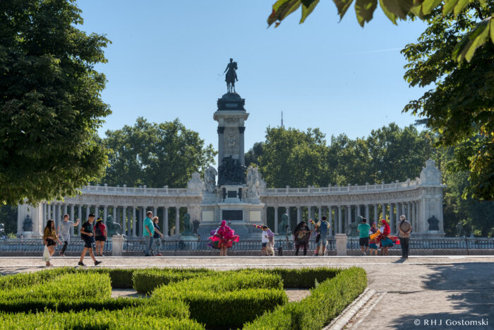Retiro Park, Madrid People walk near a large, ornate monument with a statue on top, surrounded by a semi-circular colonnade, trees, and neatly trimmed hedges in a sunny park.
