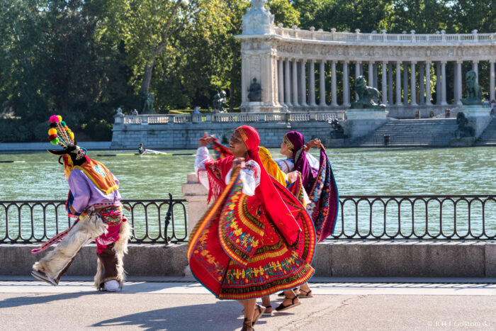 Dancing by the lake, Retiro Park, Madrid Four people in colourful traditional attire dance joyfully near a large pond with a grand colonnade and statues in the background, surrounded by trees and bathed in sunlight.