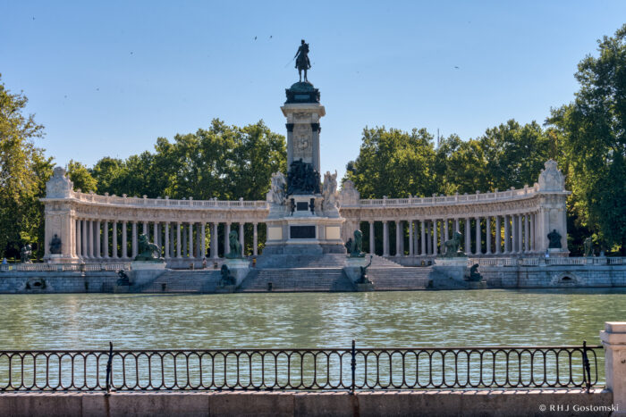 A large monument with a semicircular colonnade and a statue on top stands behind a calm lake in Parque del Retiro, with trees and a clear blue sky in the background. A black railing runs along the water’s edge in the foreground.