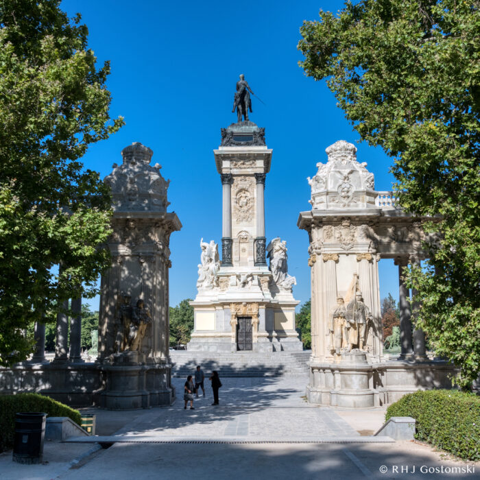 Retiro Park, Madrid A large stone monument with a statue on top stands between two ornate columns, framed by green trees under a clear blue sky. Three people are walking near the base of the steps leading up to the monument.