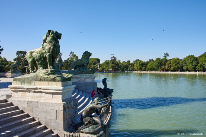 A large stone monument featuring lion statues overlooks Retiro Lake, its wide, calm waters bordered by lush green trees under a clear blue sky.