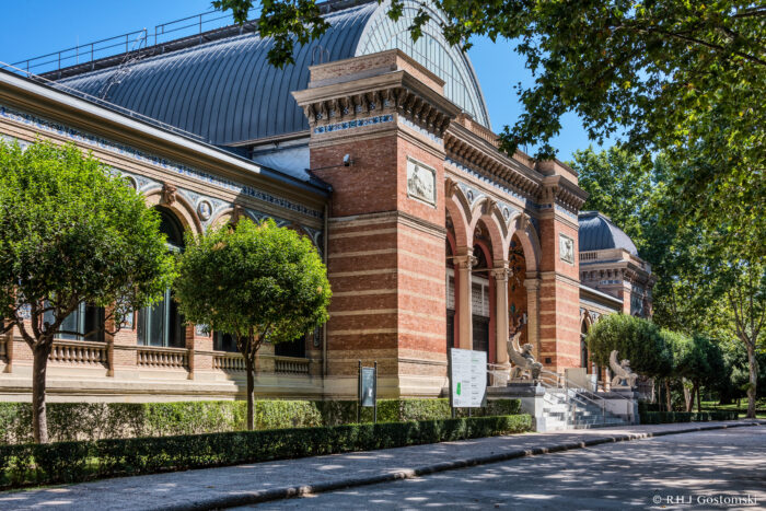 The historic brick building, Palacio de Velazquez, features arched entrances and decorative details, flanked by trimmed trees and lion statues. Sunlight and greenery enhance the scene, evoking a grand museum or cultural institution.