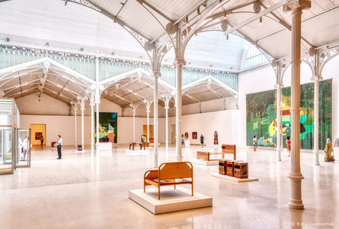 Sunny, spacious art gallery inside The Velázquez Palace in El Retiro, with high white ceilings, arched beams, large windows, and modern art pieces on display. A few visitors admire paintings and sculptures in the open, minimalist space.