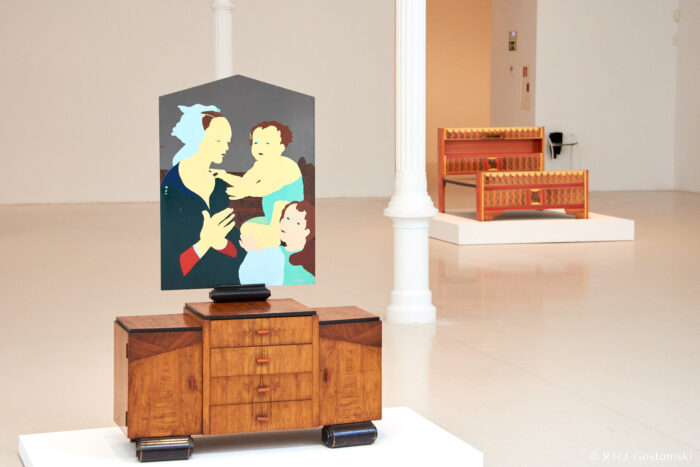Art exhibition inside The Velázquez Palace in El Retiro, Madrid A wooden cupboard with a modern painting of a woman and two children is displayed in a bright gallery, with a geometric wooden bench on a plinth in the background.