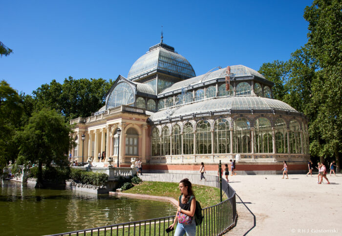 A woman strolls by a pond near the iconic Palacio de Cristal, surrounded by lush trees on a sunny day in Madrid’s Retiro Park.