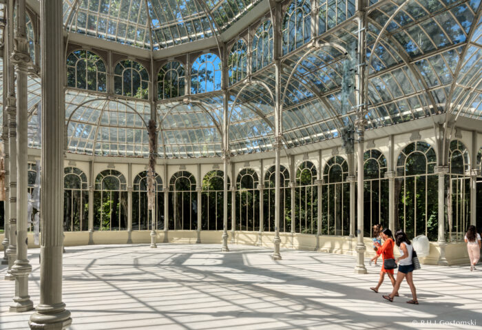 Palacio de Cristal - Madrid, Spain Three people walk inside a large glass conservatory with tall windows and intricate metal frames. Sunlight streams through the ceiling, casting shadows on the white floor. Trees are visible outside the glass walls.