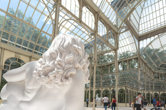 Inside the Palacio de Cristal, a large white sculpture of a reclining figure with long wavy hair is displayed, as sunlight streams through the glass ceiling and walls whilst visitors admire the stunning architecture.