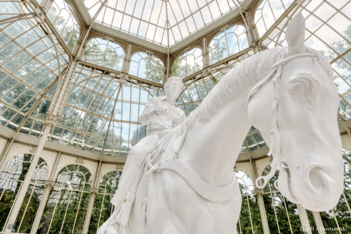 Palacio de Cristal – Madrid, Spain A white statue of a person riding a horse stands inside a glass-domed pavilion with tall windows and a metal framework. Trees and sky are visible through the glass walls and ceiling.