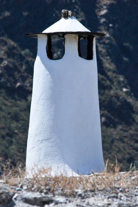 A tall, white, typical Alpujarrenian cylindrical chimney with a wide, dark-edged cap stands against a blurred mountain backdrop, with dry grass visible at the base in the foreground.