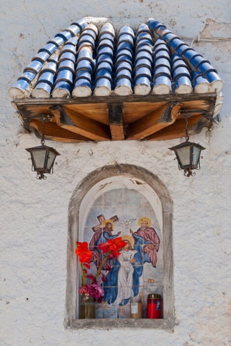 A small religious shrine set into a white wall features a painted tile image of the Virgin Mary and angels, colourful flowers, two red candles, and is sheltered by a blue-and-white tiled roof with two hanging lanterns.