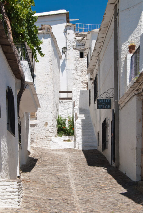 A narrow cobbled street lined with whitewashed buildings in a Mediterranean village, with bright sunlight casting shadows and a sign reading Taller Telar hanging on the right.