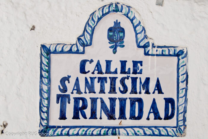 A decorative ceramic street sign with blue lettering reads Calle Sant&iacute;sima Trinidad against a white wall, bordered with a blue and white pattern and a small painted design at the top.