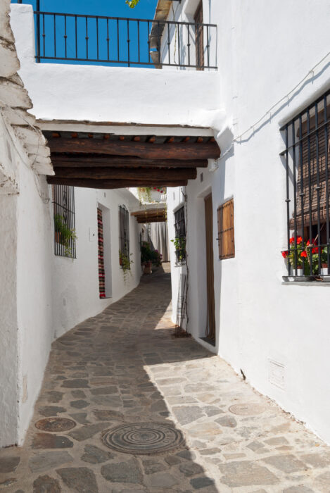 A narrow, cobbled alley with whitewashed buildings, black window grilles, and flower pots. A covered walkway connects the buildings above. The scene is bright and sunny.