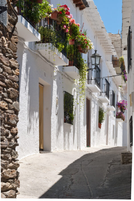 Narrow cobbled street in Capileira lined with whitewashed buildings, black iron balconies, and colourful potted flowers under a clear blue sky. Shadows from plants and railings fall across the sunlit walls and pavement.