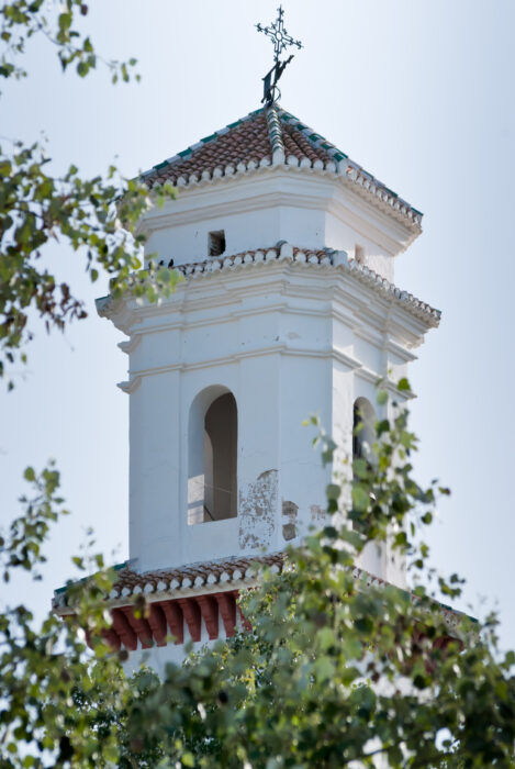 A white bell tower with arched openings and a tiled roof topped by a cross, partially obscured by green leafy branches in the foreground.