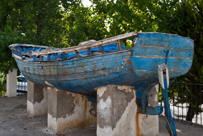 A weathered, blue wooden boat with peeling paint is displayed on concrete supports outdoors in Pitres, surrounded by lush green trees.