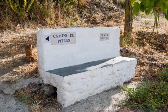A white stone bench beside a path, with two signs: one pointing left labelled Camino de Pitres and another pointing right labelled Era de Mecina. The area is surrounded by dry grass and greenery.